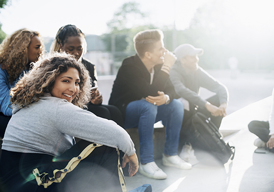 Eine Gruppe junge Leute sitzen gut gelaunt auf einem Skateplatz. Eine Frau vorne im Bild sieht lächelnd in die Kamera.