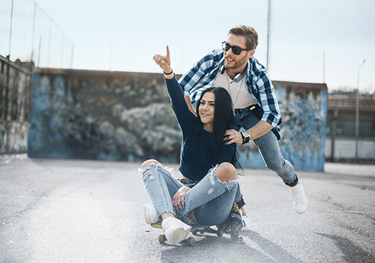 Ein junger Mann schiebt eine junge Frau, die auf einem Skateboard sitzt. Sie hält jubelnd die Hand hoch.